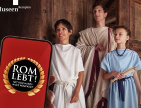 Drei als Römer der Antike gewandete Personen - ein Erwachsener und ein Junge und ein Mädchen- posieren in einer geöffneten Holztüre für ein Handy, das im Vordergrund sichtbar ist und den Namen der Ausstellung auf dem Display trägt